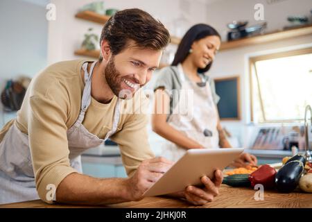 Primo piano del giovane felice che naviga sul tablet digitale alla ricerca di una ricetta da seguire online mentre cucinava a casa con sua moglie Foto Stock