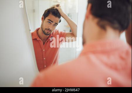 Bel giovane caucasico che tocca i capelli e guarda nello specchio del bagno. Maschio tirando i capelli e pensando di ottenere un taglio di capelli. Interessato Foto Stock