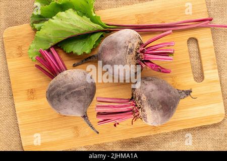 Tre barbabietole rosse dolci con un tagliere in legno sul burlap, primo piano, vista dall'alto. Foto Stock