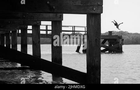 Boy Jumping dal Wooden Pier sulla spiaggia di Lake Pier sulla costa settentrionale di Poole Harbour, Regno Unito Foto Stock