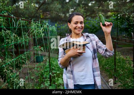 Affascinante donna multietnica agricoltore dilettante, giardiniere che detiene un cetriolo fresco raccolto maturo coltivato nella sua fattoria ecologica di famiglia. Il futuro è vegano. Amore e. Foto Stock