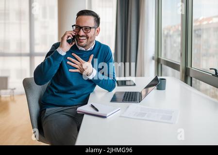 Felice uomo d'affari millenario che effettua una telefonata al cliente, consulti il cliente, parla sullo smartphone davanti al computer portatile in ufficio w Foto Stock