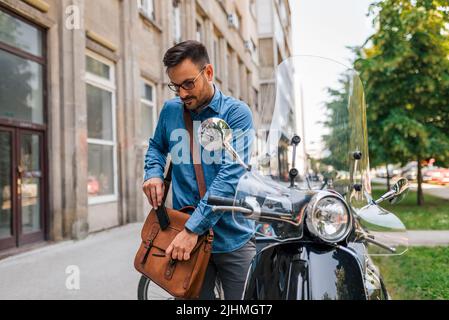 Sorridente uomo d'affari che chiude la borsa mentre si prepara a guidare il suo scooter a motore. Professionista di sesso maschile in piedi da un veicolo parcheggiato sul marciapiede. È in viaggio Foto Stock