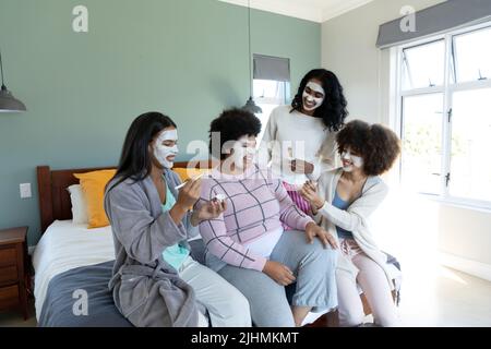 Allegro biraciale giovani amiche con creme di bellezza sui volti che trascorrono tempo libero a letto Foto Stock