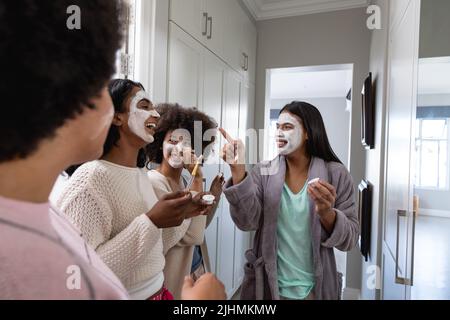 Allegro biraciale giovani amiche con creme di bellezza sui volti che trascorrono tempo libero a casa Foto Stock
