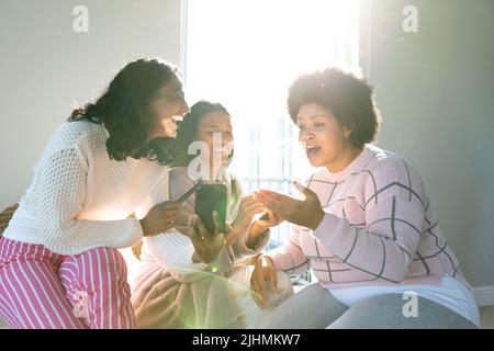 Felice giovane donna biraciale che mostra smartphone a un amico femmina ridendo mentre si siede dalla finestra Foto Stock