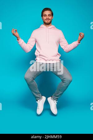 Tutta la lunghezza di un uomo attento e attento che si siede facendo yoga mentre salta isolato su sfondo di colore blu chiaro Foto Stock