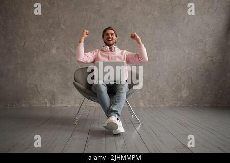 Maschio gioioso usando il laptop e scuotendo i Fists che festeggiano le grandi notizie e la fortuna seduta nella sedia sopra sfondo grigio della parete Foto Stock