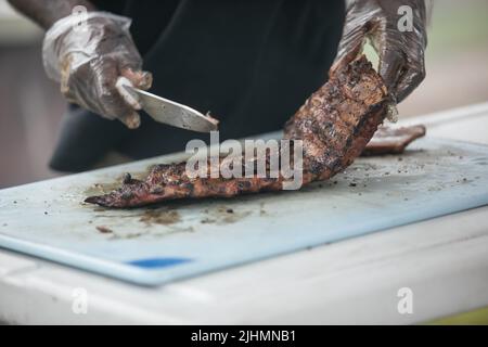 Un uomo che taglia le costole del barbecue dalla griglia fumatore sul tagliere con i guanti sopra e un coltello grande. Foto Stock