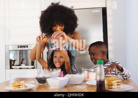 Madre multirazziale con capelli afro rompendo l'uovo con la figlia in ciotola mentre fa la colazione sul tavolo Foto Stock
