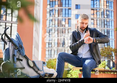 Fai una chiamata d'affari, guardando l'orologio mentre trascorre del tempo con figlio all'aperto. Vista frontale dell'uomo bearded che tiene il telefono mobile, che chiama, mentre il bambino dorme in pram nel parco. Concetto di lavoro. Foto Stock