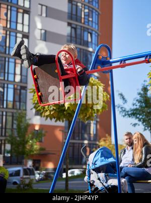 Piccolo figlio che si diverte in altalena, mentre mamma e papà seduti su panchina con il pam al parco giochi. Vista laterale del ragazzo gioioso sul swing, mentre i genitori si stanno cura del bambino dietro al giorno. Concetto di famiglia. Foto Stock