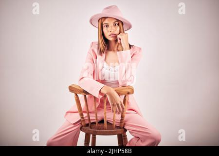 Ritratto di giovane donna seria gara mista in posa in abiti alla moda, seduto su una sedia in uno sfondo grigio studio. Donna ispanica Foto Stock
