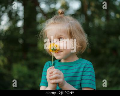 Ritratto di primo piano di un ragazzino con capelli biondi lunghi che trattengono e odorano fiori con petali gialli all'esterno Foto Stock