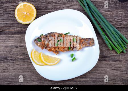 Pesce fritto impanato con cipolle verdi e limone, in un piatto su un vecchio sfondo di legno Foto Stock