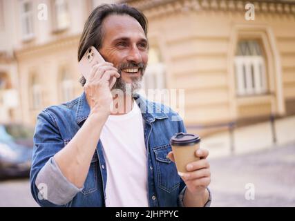 Felice uomo di mezza età con portico grigio parlare sul telefono tenendo il caffè in carta usa e getta tazza in piedi all'aperto nella città vecchia sfondo indossare jeans camicia. Uomo in viaggio freelance. Foto Stock