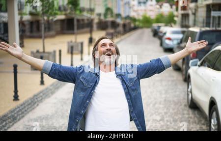 Felice uomo anziano grazie a Dio in piedi all'aperto con le mani spalancate ampia camicia da indossare jeans con t-shirt bianca sotto. Uomo di mezza età contento che la vita è bella in piedi nella strada della città vecchia. Foto Stock