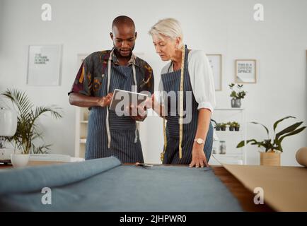 Due progettisti di abbigliamento che utilizzano un tablet digitale mentre lavorano con il materiale sul lavoro. Giovane afroamericano su misura che parla e tiene un tablet digitale Foto Stock