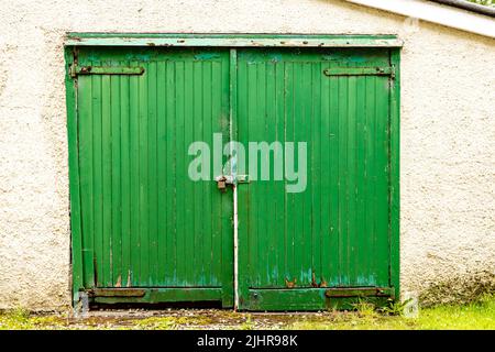 Dritto in vista di un paio di porte da garage verde lucchettate che hanno visto giorni migliori con cerniere a cinghia, vernice peeling e legno marcio Foto Stock