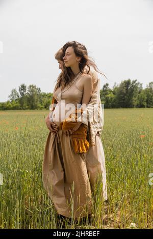 Vista laterale dell'uomo che abbraccia la moglie incinta sul campo Foto Stock