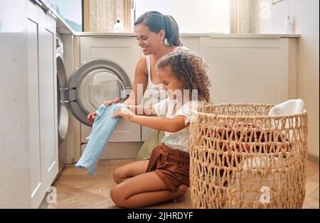 La giovane madre ispanica e sua figlia che smistano la biancheria sporca nella lavatrice a casa. Adorabile bambina e sua madre che fanno i compiti insieme Foto Stock