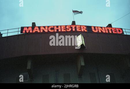 1990s Vista esterna dello stadio Old Trafford, sede del Manchester United Football Club, Manchester, Inghilterra, Regno Unito. Cartello rosso al neon con il nome del club in cima all'East Stand. Un orologio qui sotto ricorda un momento decisivo nella storia del club, il tragico disastro di Monaco della data di febbraio 6th 1968, quando molti dei più grandi giocatori e funzionari del club sono morti come il loro aereo si è schiantato in una bufera sulla pista aeroportuale della città. Foto Stock