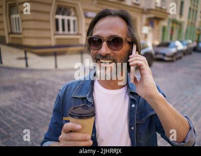 Primo piano ritratto parlando al telefono uomo medio vecchio bearded con take away caffè in carta tazza in piedi all'aperto nella città vecchia indossando jeans camicia. Uomo in viaggio freelance. Lavoro in movimento. Foto Stock