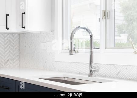 Un dettaglio lavandino della cucina girato con mobili blu e bianchi, backsplash in piastrelle a spina di pesce e un rubinetto cromato di fronte a una finestra. Foto Stock
