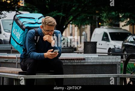 Un uomo con uno zaino Wolt blu per la consegna di cibo seduto su una panca e guardando il suo smartphone Foto Stock