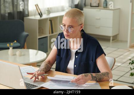 Donna economica seria in occhiali che lavorano sul portatile a casa per pagare le fatture e le tasse sul gadget online Foto Stock