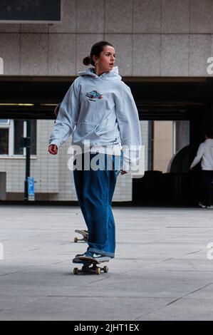 Un colpo verticale di una bella giovane donna in una felpa con cappuccio grigia che guida uno skateboard per le strade di Praga Foto Stock