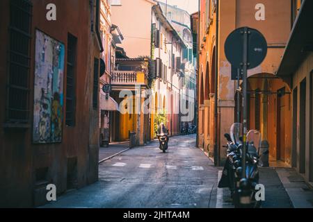 Un uomo in scooter che percorre una stradina stretta con edifici colorati a Bologna, in Italia di giorno Foto Stock