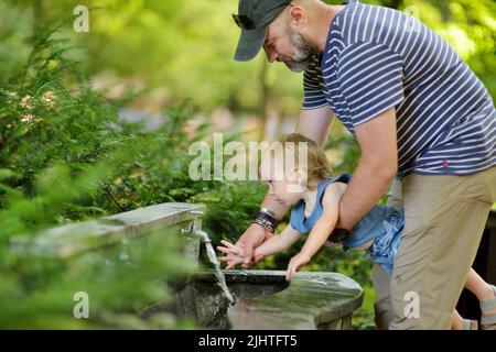 Padre che aiuta il figlio del bambino a lavarsi la mano nella fontana della città. Papà e figlio che si divertono nella soleggiata giornata estiva della città. Foto Stock