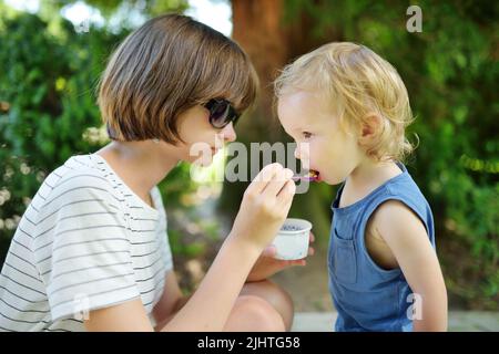Bella ragazza adolescente mangiare gustoso gocce gelato all'aperto nella soleggiata giornata estiva. Sorella grande che alimenta un dessert al fratello del bambino. Dolci per bambini. Foto Stock
