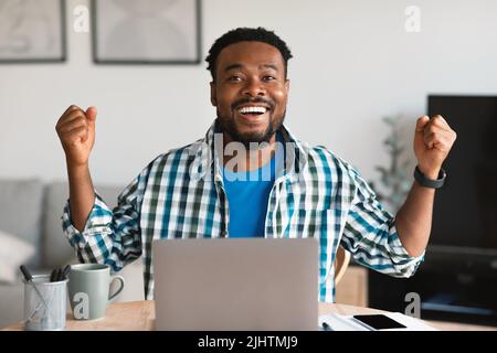 Joyful Black Male utilizzando il computer portatile gesturing Sì a casa Foto Stock