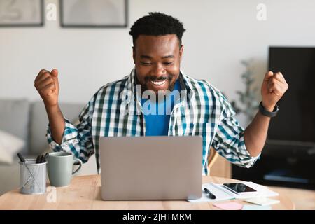 Gaudioso ragazzo afroamericano che usa il laptop scuotendo i fisti seduti al chiuso Foto Stock