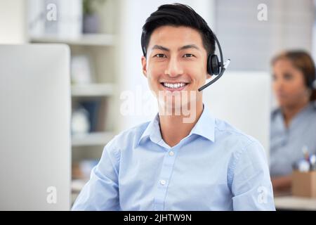 Ritratto dell'agente del call center asiatico che parla con le cuffie mentre lavora con il computer in ufficio. Fidato e sorridente uomo d'affari consulenza e funzionamento Foto Stock