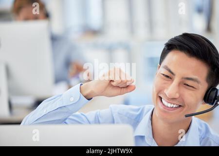 Un uomo d'affari asiatico eccitato festeggia un successo acclamando e punzonando l'aria con i suoi pugni mentre si siede a una scrivania. Un agente del call center con cuffia Foto Stock