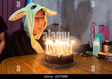Un ragazzo soffia le candele su una torta di compleanno al cioccolato mentre indossa un abito da condimento Yoda. Foto Stock