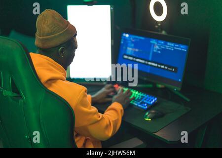 Vista posteriore di un afroamericano che indossa una felpa con cappuccio arancione e un cappello seduti su una sedia alta, guardando uno schermo del computer. . Foto di alta qualità Foto Stock