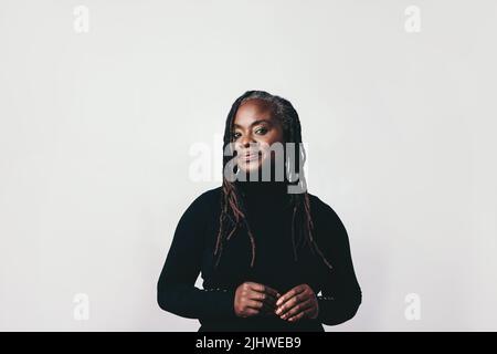 Donna di mezza età sicura con dreadlock che guarda la fotocamera in uno studio. Elegante donna nera in piedi su uno sfondo grigio. Foto Stock