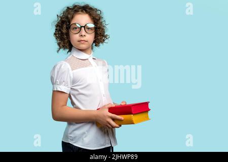 La studentessa curly con taglio corto in blusa bianca e occhialini rotondi contiene libri di testo su sfondo blu studio. Libri rossi e gialli in mani di g Foto Stock