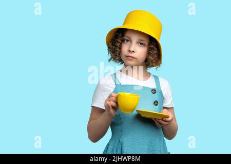 Сurly Schoolgirl a capelli corti 10-12s in cappello panama giallo e Sundress blu estate tenendo una tazza gialla con piattino su sfondo studio blu chiaro. Gir Foto Stock