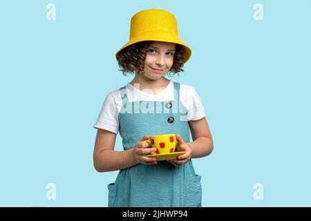 Festa del tè. Сurly ragazza dai capelli corti 10-12s in cappello panama giallo e spogliatore che tiene una tazza gialla con piattino su sfondo studio blu chiaro. Elega Foto Stock