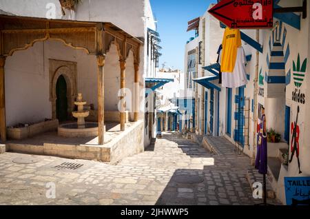Una corsia fiancheggiata da tradizionali edifici dipinti di blu e bianco conduce giù per la collina, passando davanti ad una fontana nell'antica Medina di Sousse in Tunisia. Foto Stock