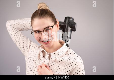 Donna sorridente con bretelle usa una pistola massaggiatore per la schiena. Foto Stock