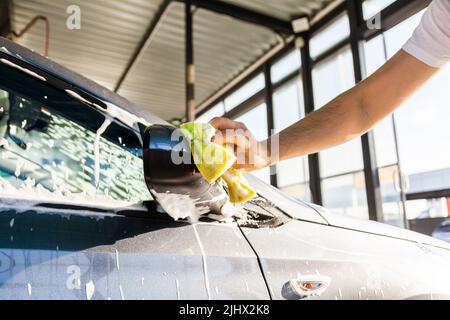 Un uomo lava lo specchio sulla sua auto con schiuma ad un autolavaggio auto self-service, primo piano. Foto Stock