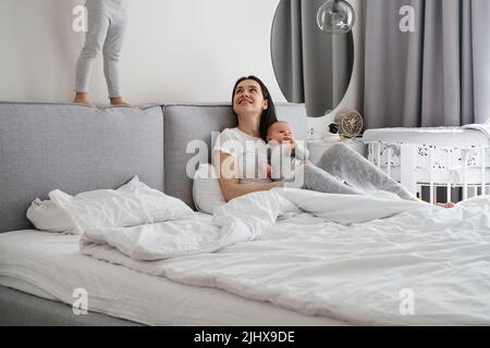 Mama con due bambini a casa in camera da letto. Bambino bambino bambino che riposa sulle mani delle madri, mentre la sorella più anziana che gioca, azione, saltando sul letto vicino, Cuddle A. Foto Stock