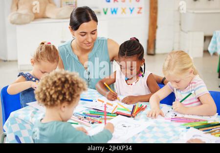 Bella donna gara mista accovacciata giù per guardare un gruppo di diversi bambini colore in prescolare. Bambini piccoli e carini seduti insieme e disegno Foto Stock