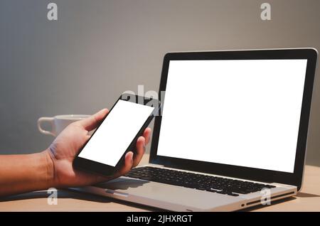 man hand holding mobile smartphone and computer laptop mock up blank screen on desk. Foto Stock